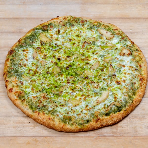 A whole pesto chicken pizza with melted cheese and a golden crust on a wooden surface.