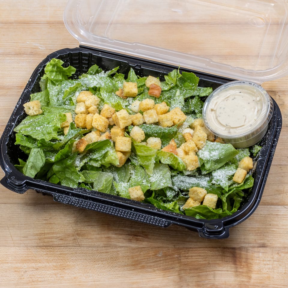 Caesar salad with croutons and dressing in a black plastic container on a wooden surface.
