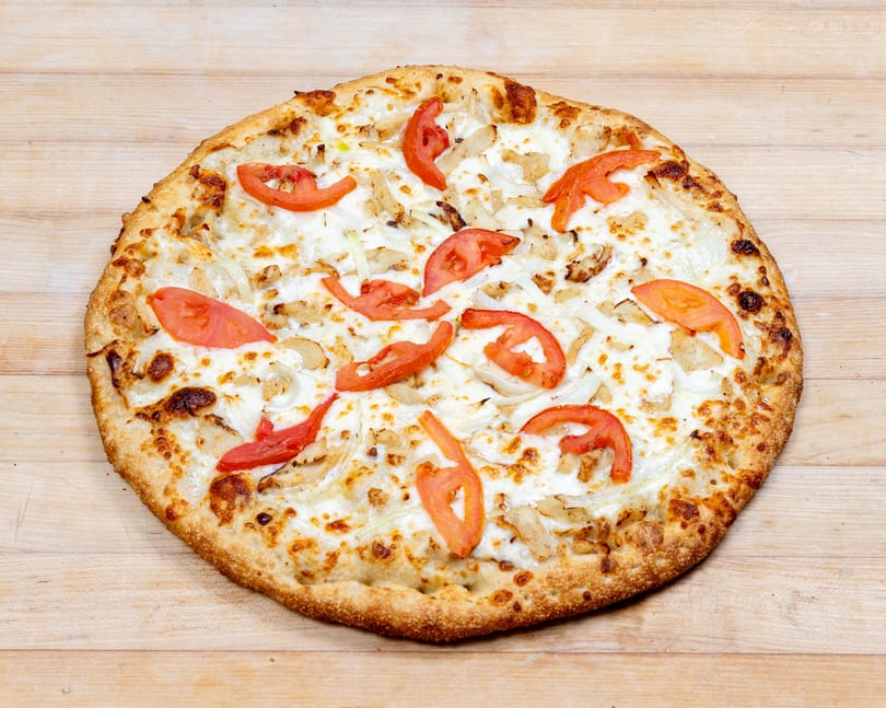 Round pizza with white sauce, melted cheese, chicken, onions, and tomato slices on a wooden surface.