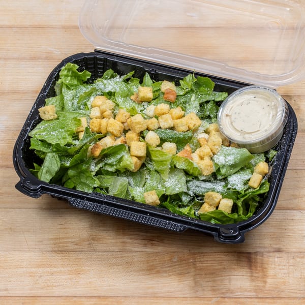 Caesar salad with croutons and dressing in a black plastic takeout container on a wooden surface.