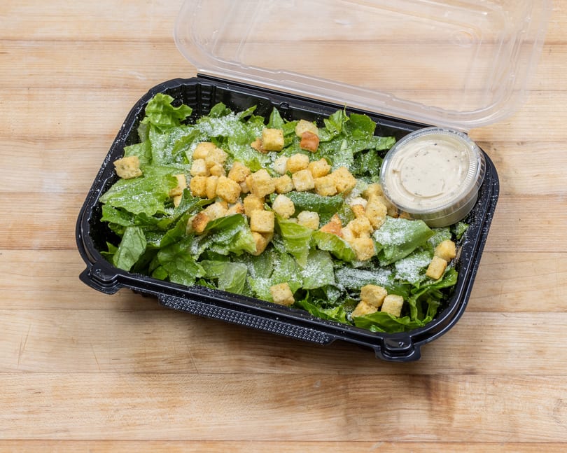 Caesar salad with croutons and dressing in a black plastic takeout container on a wooden surface.