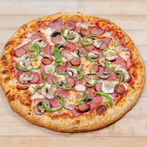 A whole pizza with sausage, mushrooms, green peppers, and ham on a light wooden surface.