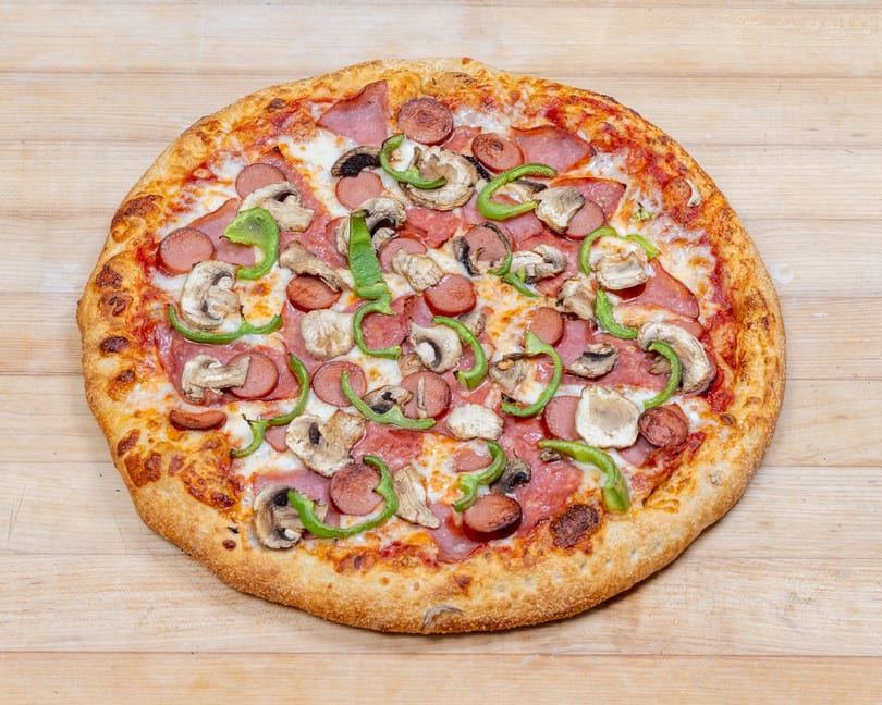 A whole pizza with sausage, mushrooms, green peppers, and ham on a light wooden surface.