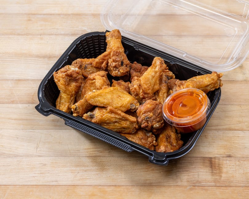 Crispy fried chicken wings and a small container of hot sauce in a black to-go box.