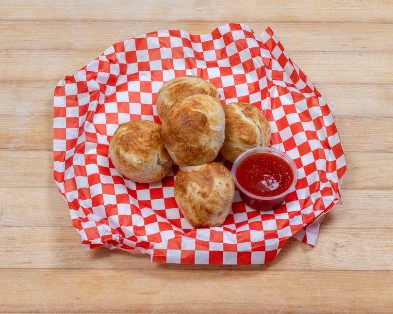 Five golden-brown baked dough balls and a container of red sauce on red and white checkered paper.